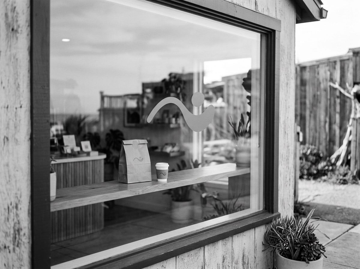 Café window with a large brand mark, with a branded paper bag and cup on the counter inside
