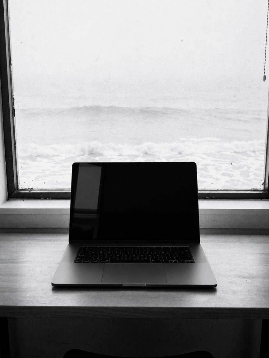 Laptop on a wooden counter in front of a window looking out on breaking waves